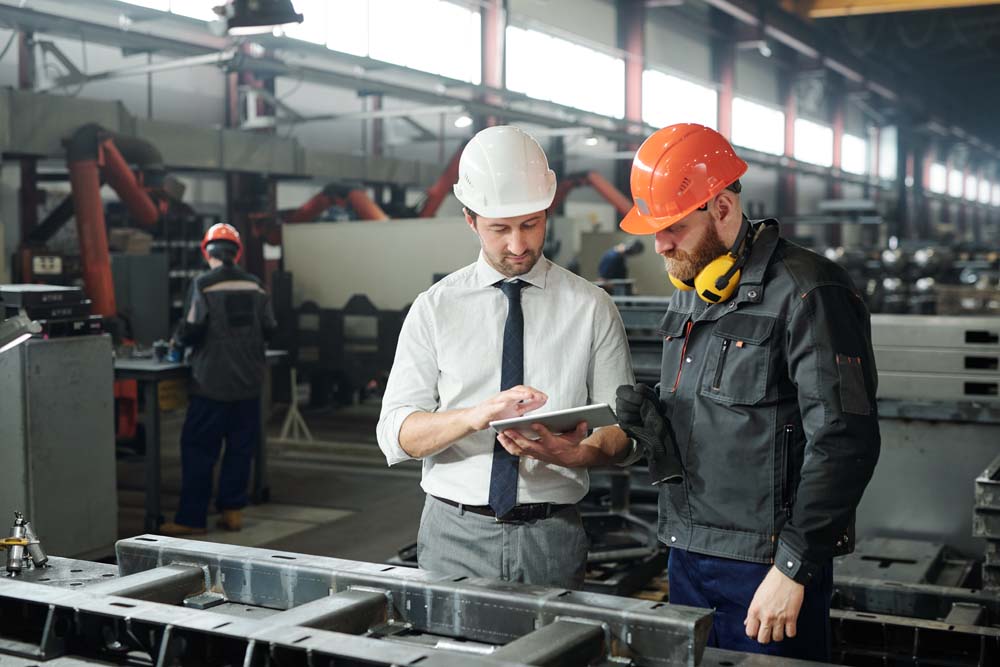 Young leader in hardhat and bearded engineer discussing technical sketch on display of tablet in factory workshop