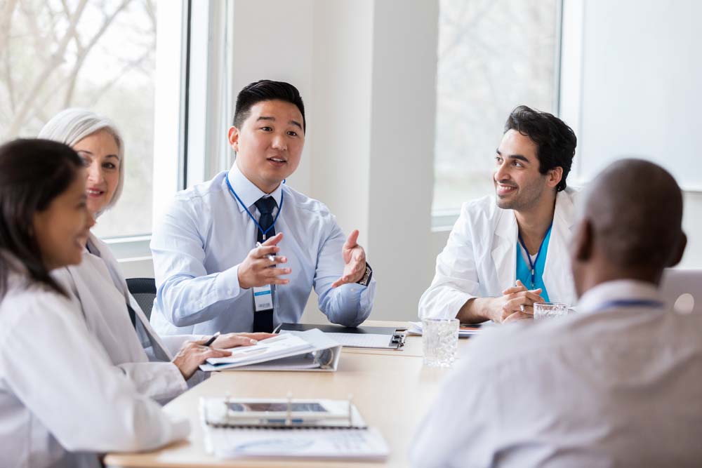 Young male hospital administrator gestures while discussing data that was generated by BigBear.ai AI software with doctors and other people in hospital management.