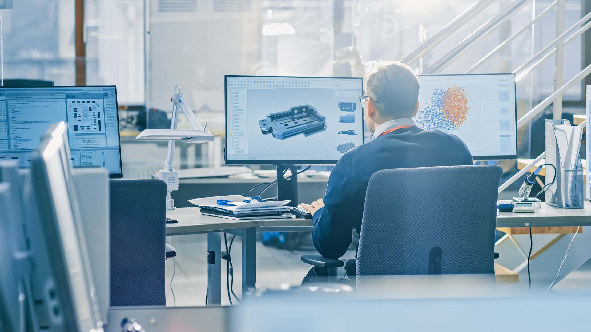 Back View of Industrial Engineer Working on Desktop Computer in bright office. Screens Show IDE / CAD Software, Implementation of Machine Learning, Neural Networking and Cloud Computing