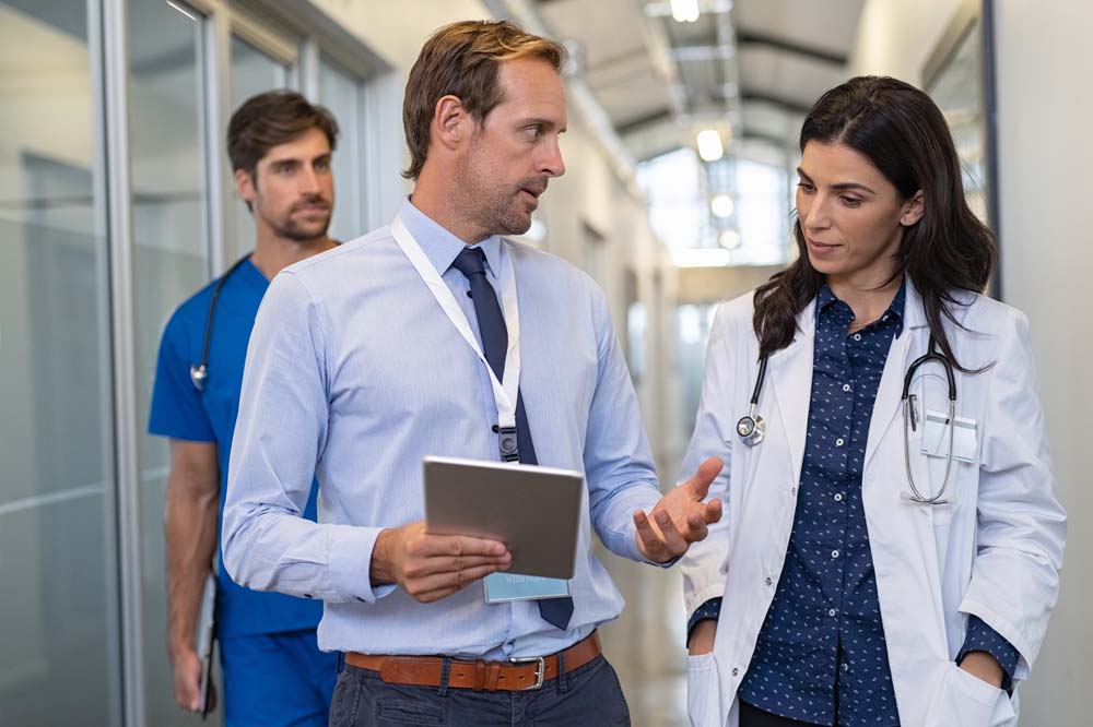 Man and woman doctor having a discussion in hospital hallway while holding digital tablet equipped with BigBear.ai software. The pharmaceutical representative shows a medical report that was analyzed and generated by AI software