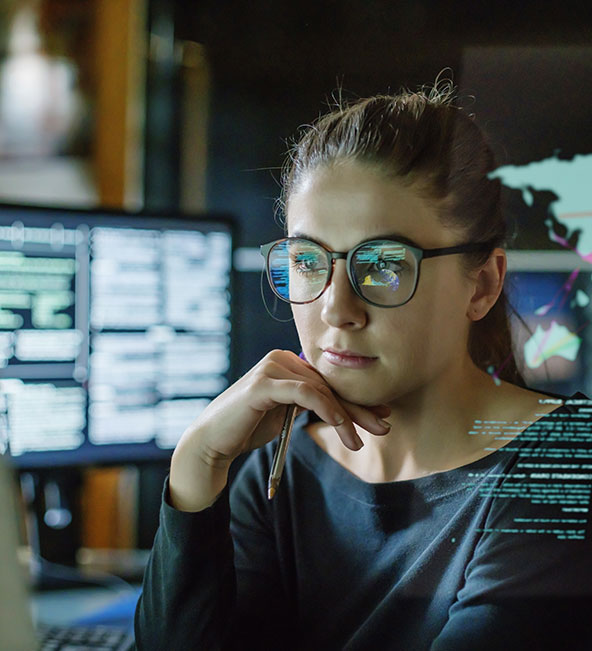 young woman, wearing glasses, surrounded by computer monitors in a dark office. In front of her there is a see-through displaying showing a map of the world with some data.
